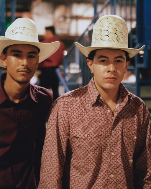 Two young adult males wearing cowboy hats and button-down shirts stand together in front of metal fencing and blurry figures, medium shot.