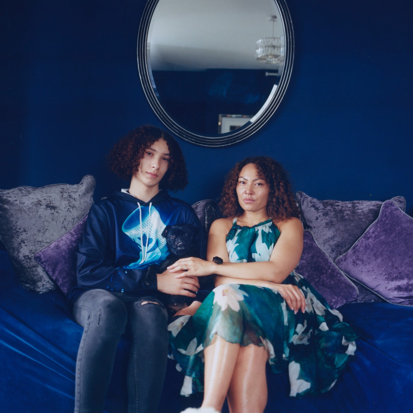 Adult woman and teenager sit on a blue couch with a small dog. The background is a dark blue wall with an oval mirror.