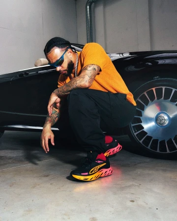 Stylish man in yellow shirt and black pants crouching next to a modern black car with chrome details, wearing sunglasses and sneakers.