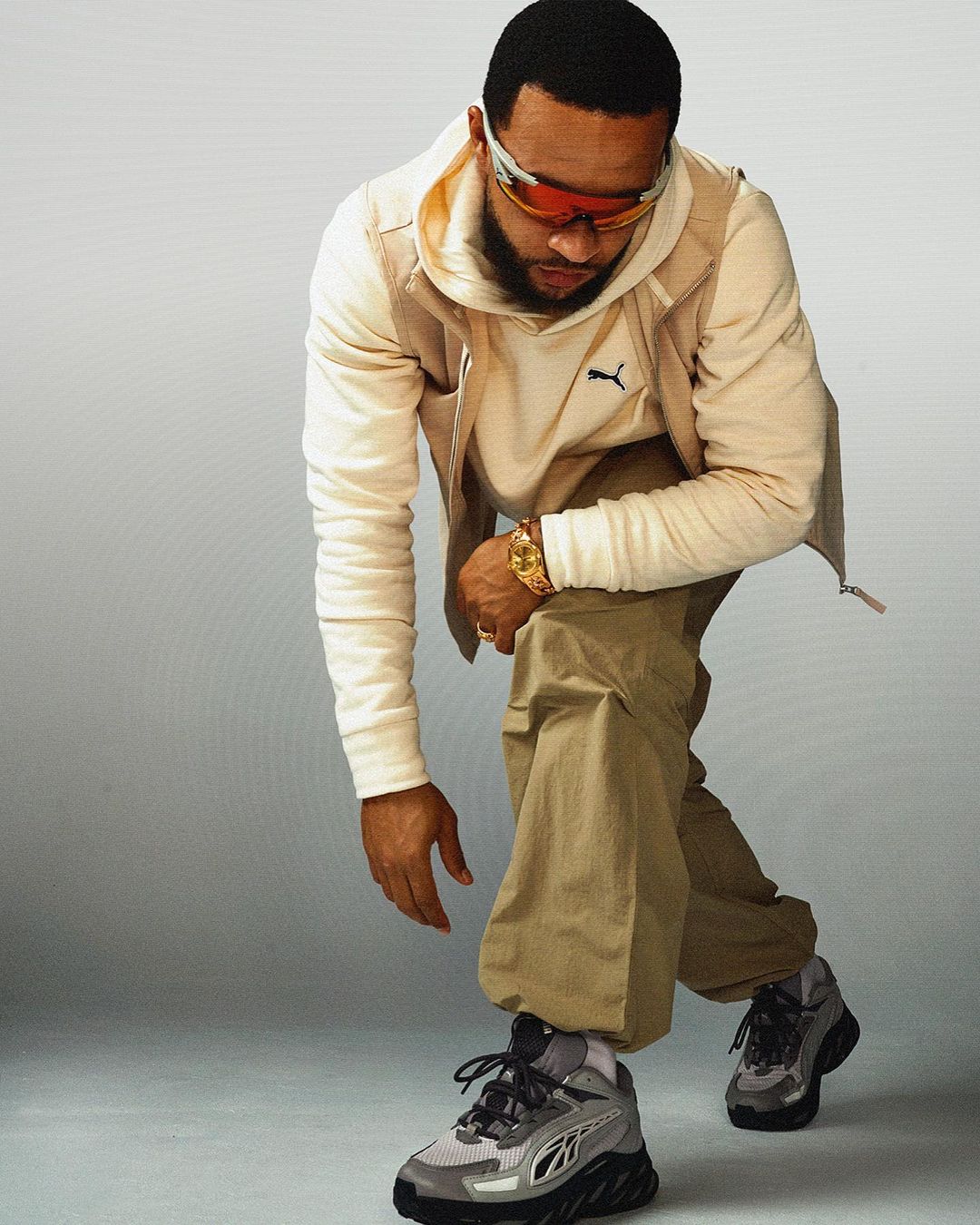 Man in a cream hoodie and cargo pants poses in a photo studio. He wears orange goggles and black and grey sneakers against a light grey backdrop.