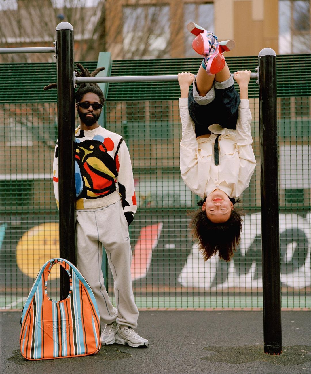 Child hanging upside down on a bar with an adult and tote bag nearby in an urban outdoor setting on a sunny day.