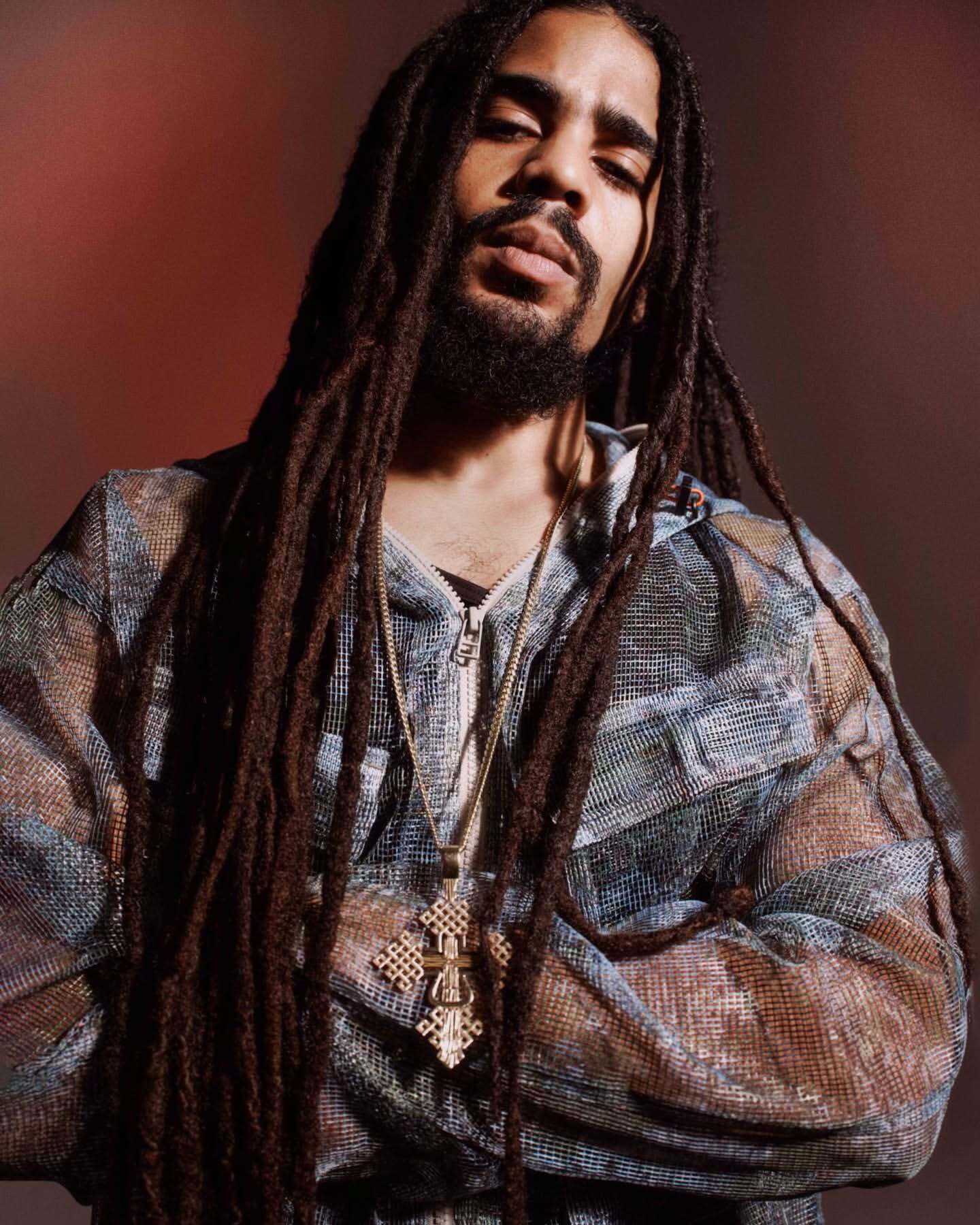 Portrait of a man with long dreadlocks wearing a gold cross necklace and a patterned hooded jacket against a blurred backdrop.