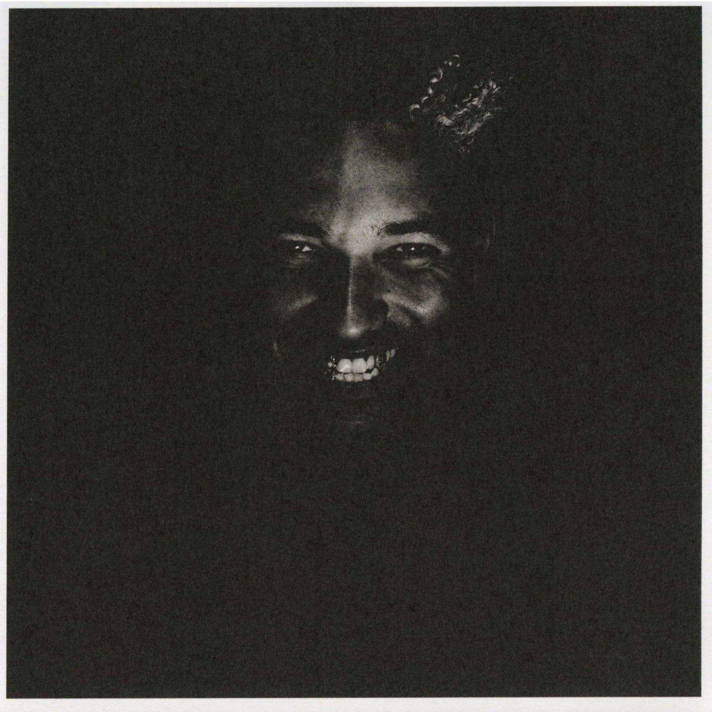 Close-up portrait of an adult with curly hair smiling broadly in deep shadow, with only the person's face and upper chest visible.