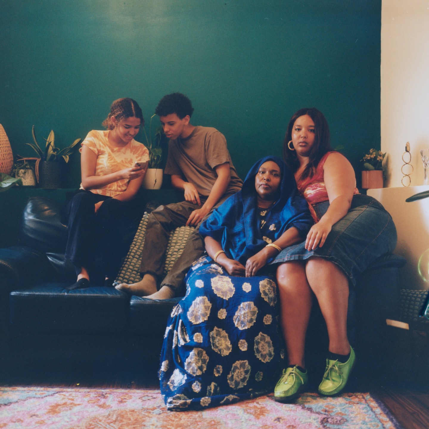 A group of four people of mixed ages and ethnicities are sitting on a couch in an indoor setting with plants and a green wall.