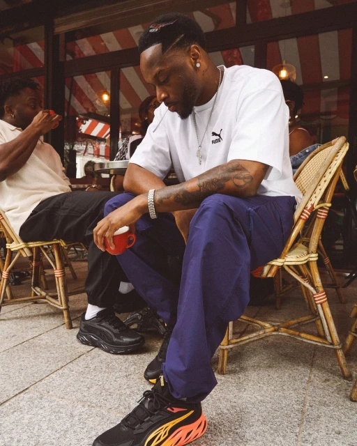 A dark-haired adult male, wearing a white Puma t-shirt and black and orange Puma sneakers, sits on a chair at an outdoor cafe.