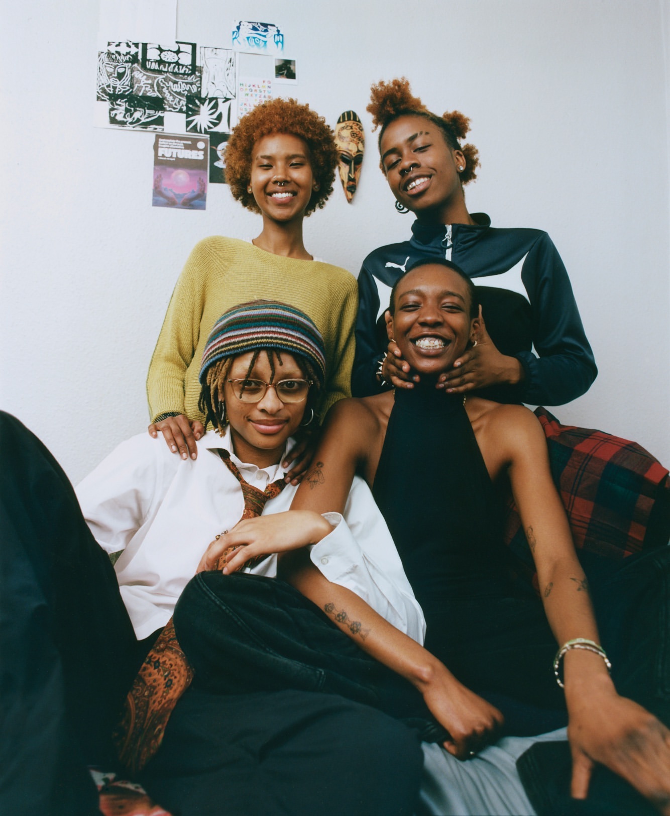 Group portrait of four young Black women smiling indoors. They wear a mix of casual clothing, with decorative elements on the wall behind them.