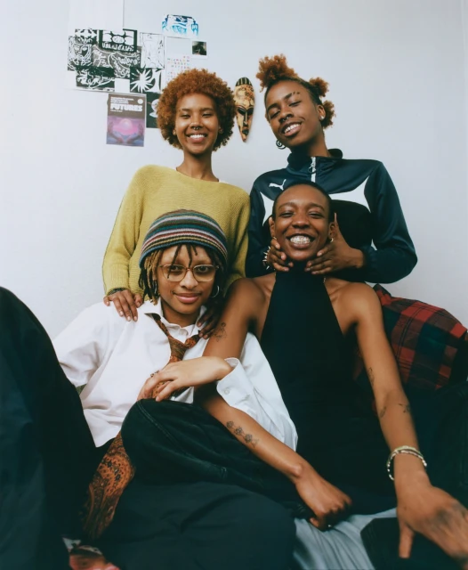 Group portrait of four young Black women smiling indoors. They wear a mix of casual clothing, with decorative elements on the wall behind them.