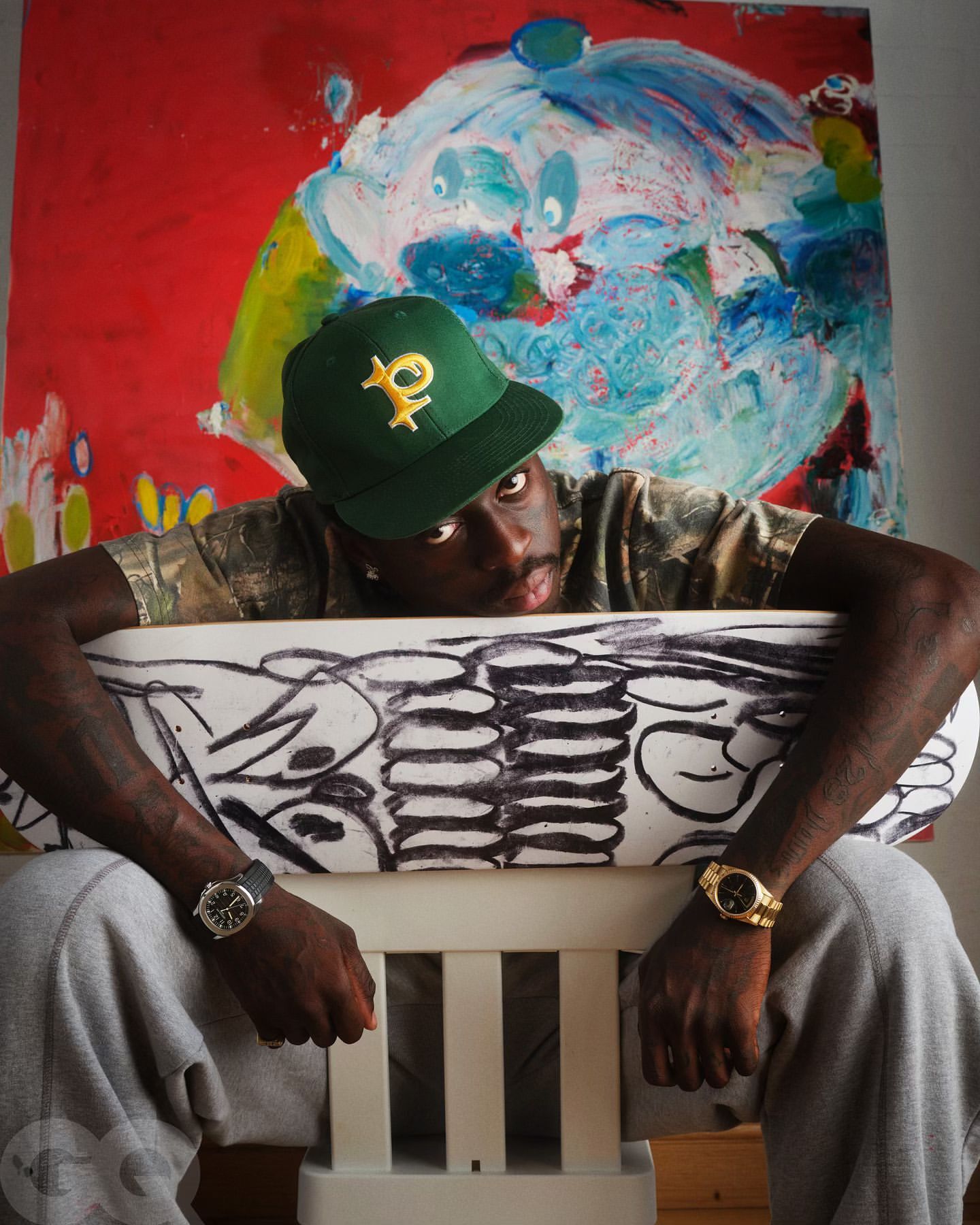 Person seated with hands on a drawing, wearing a green baseball cap, wristwatch, gray sweatpants; colorful painting in background.