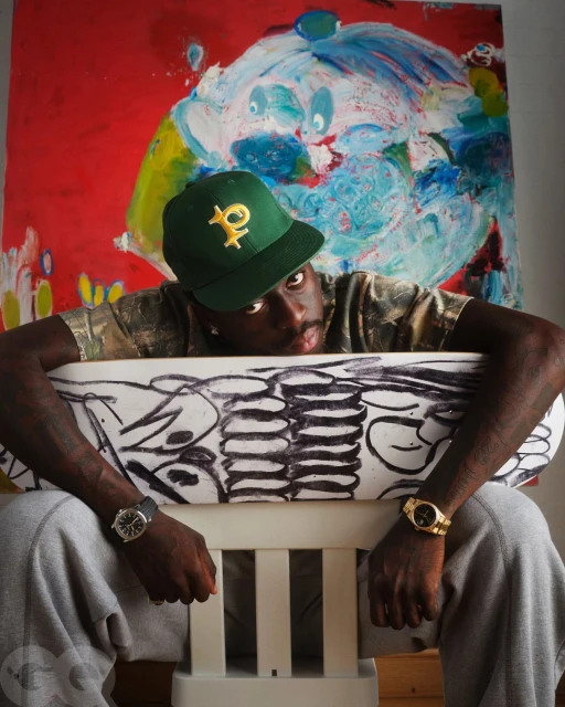 Person seated with hands on a drawing, wearing a green baseball cap, wristwatch, gray sweatpants; colorful painting in background.