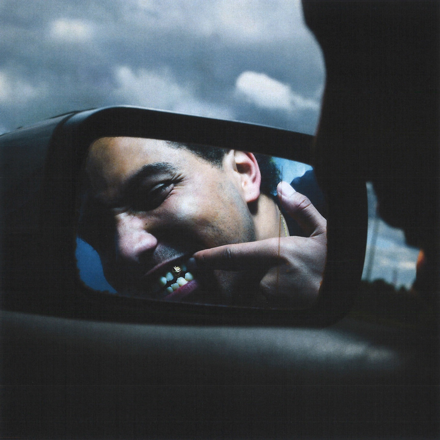 Man looks in a side mirror displaying gold teeth and pointing at them with his finger, outside on an overcast day.