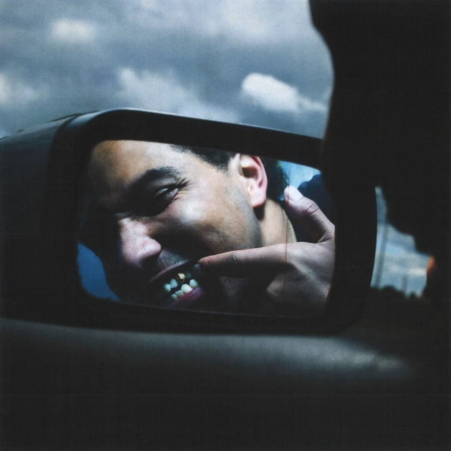 Man looks in a side mirror displaying gold teeth and pointing at them with his finger, outside on an overcast day.