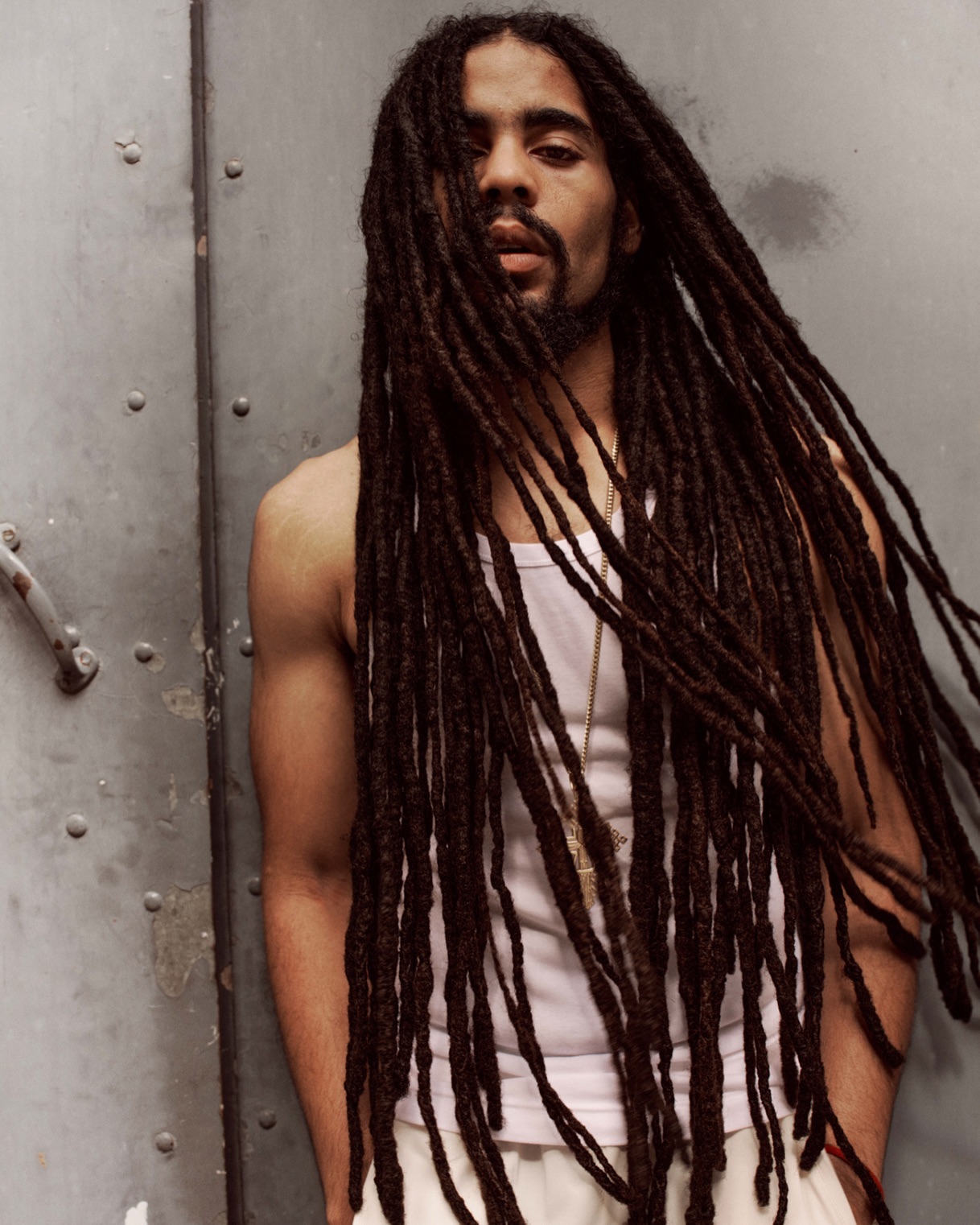 Portrait of a Black adult male with long dreadlocks, wearing a tank top and pants, posing against a gray wall, medium shot.