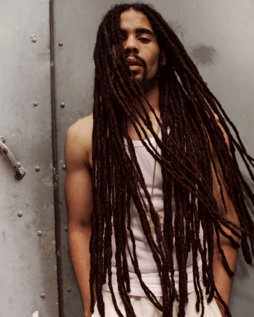 Portrait of a Black adult male with long dreadlocks, wearing a tank top and pants, posing against a gray wall, medium shot.