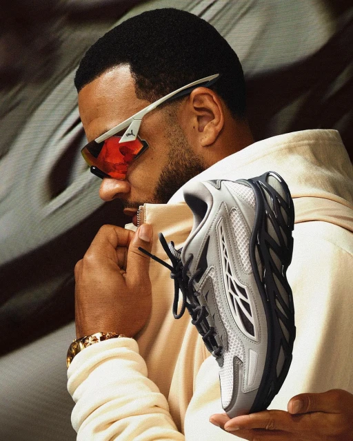 Man holding gray athletic shoe, wearing red and white sunglasses and gold bracelet, tan sweatshirt, indoor setting, soft lighting.