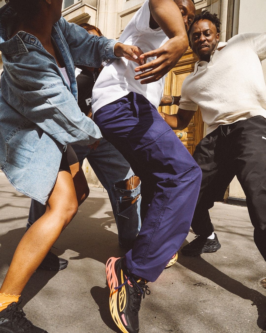 A group of four African-descent young adults gather outside a building, two dancing and wearing streetwear fashions.