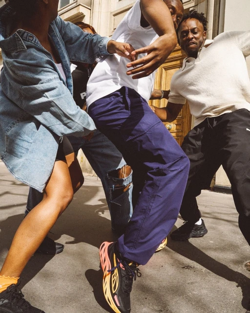 A group of four African-descent young adults gather outside a building, two dancing and wearing streetwear fashions.