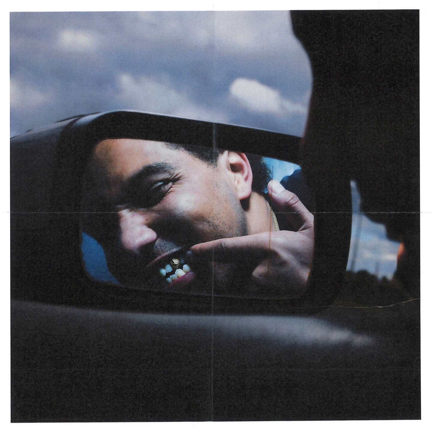 Reflection in car mirror of a smiling adult male pointing to his gold teeth, cloudy sky background.
