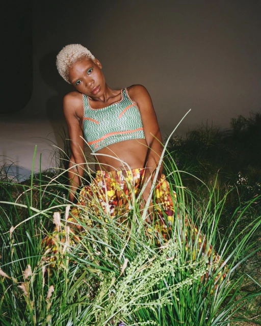 Woman with short bleached hair poses in tall grass, wearing a green geometric patterned top and floral skirt against a grey backdrop.
