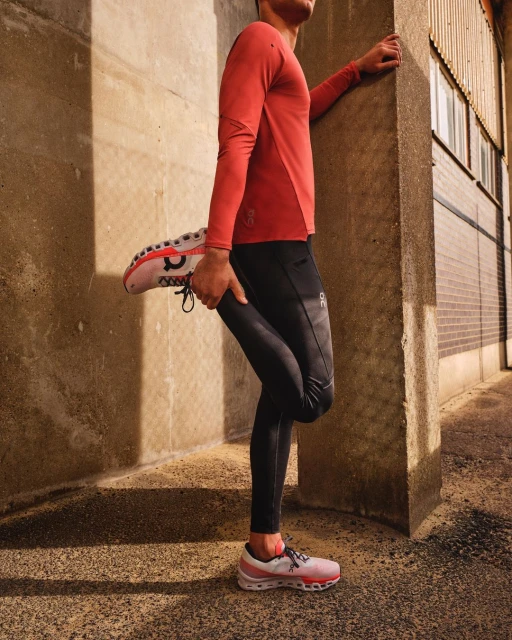 Male athlete stretches leg against a concrete pillar outdoors in natural light wearing athletic top and leggings with running shoes.