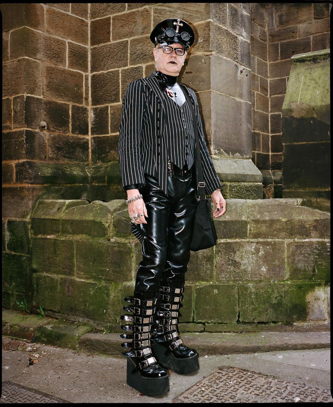 An adult wearing gothic fashion stands outdoors against a brick wall in platform boots, pinstripes, and a black cap with goggles.