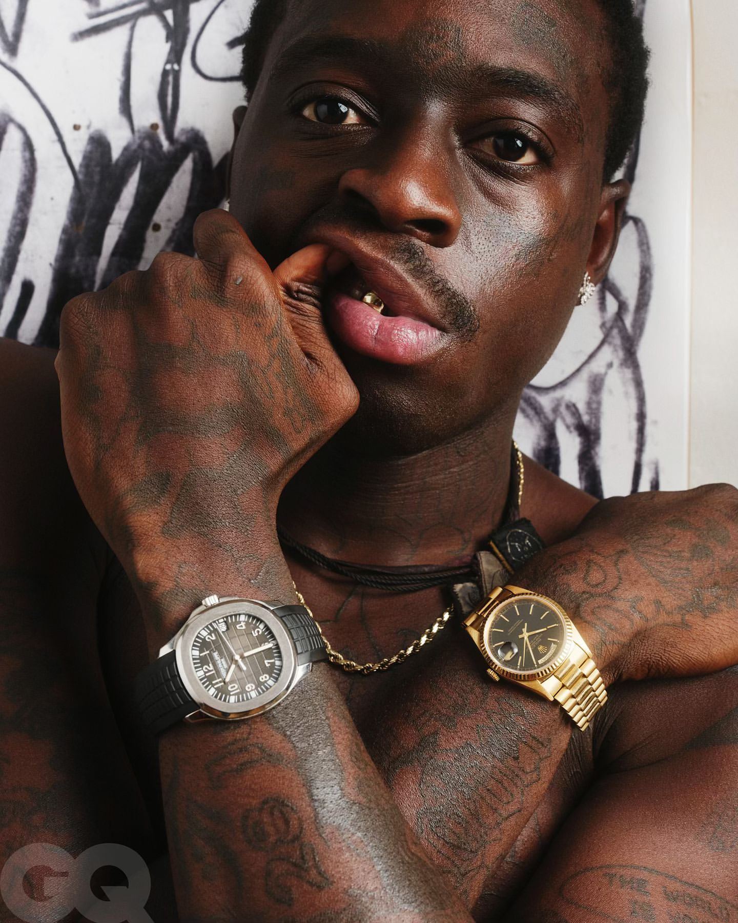 Close-up portrait of a Black adult male with gold teeth and multiple watches, wearing necklaces against a graffiti background.