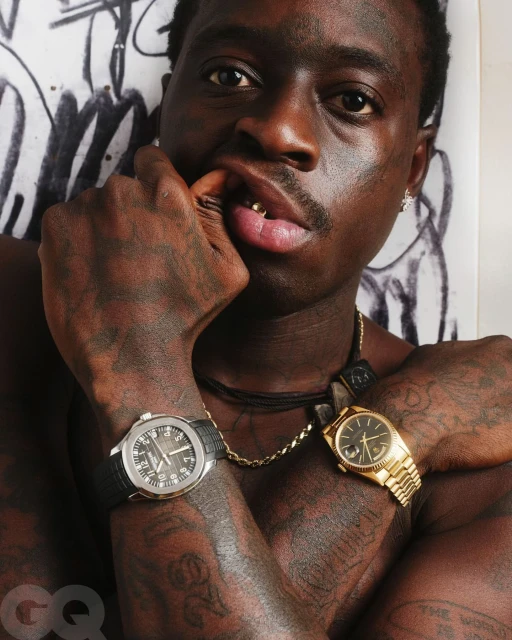 Close-up portrait of a Black adult male with gold teeth and multiple watches, wearing necklaces against a graffiti background.