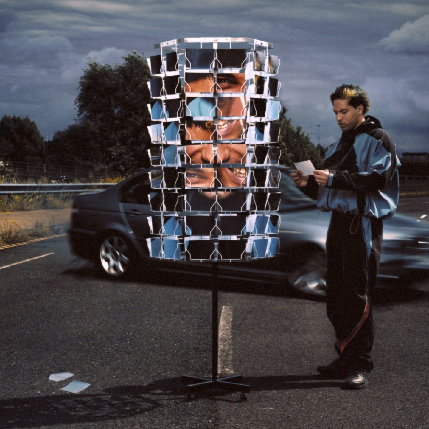 Man views postcards on a revolving stand along a highway, with a car passing by and cloudy sky overhead in a conceptual outdoor scene.