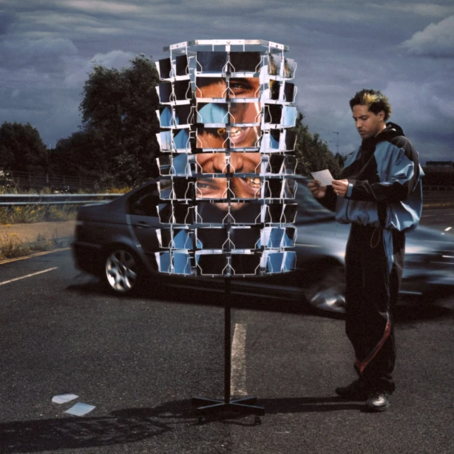 Man views postcards on a revolving stand along a highway, with a car passing by and cloudy sky overhead in a conceptual outdoor scene.