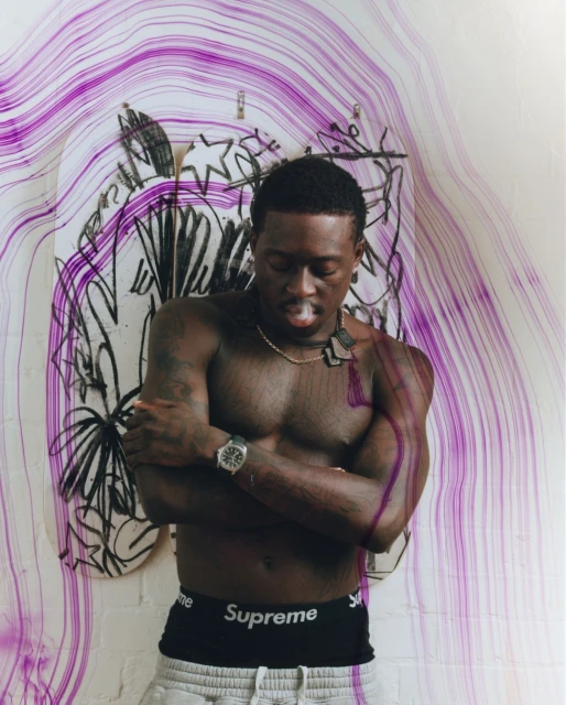 Shirtless Black man with watch poses in front of painted skateboards and purple light trails in an indoor setting, casual style.