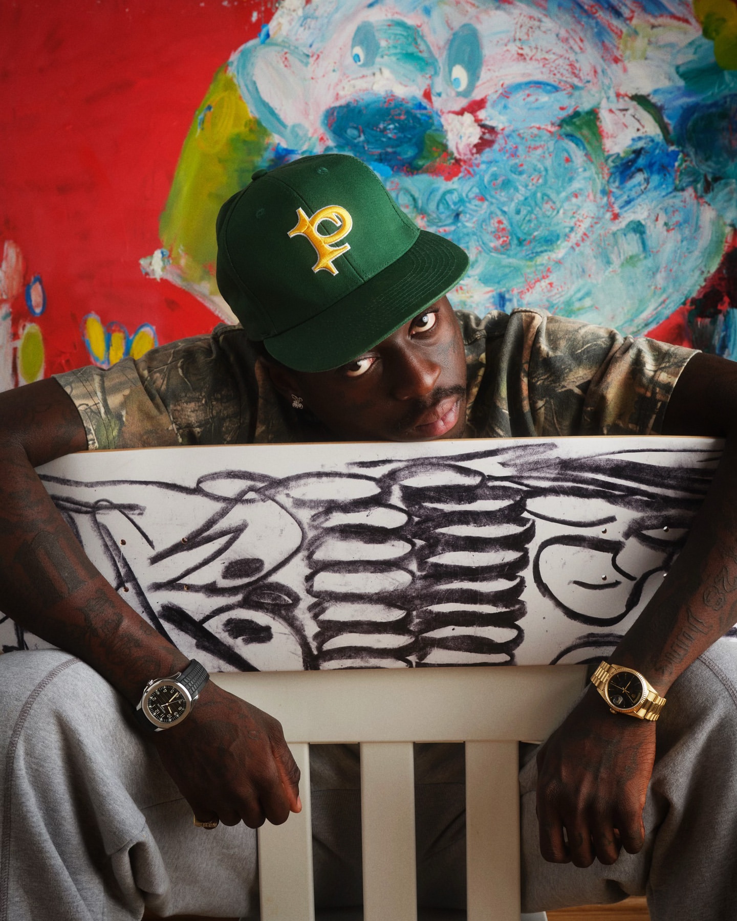 A man wearing a green cap and two wristwatches leans on a white surface with a black graphic design, against a colourful background.