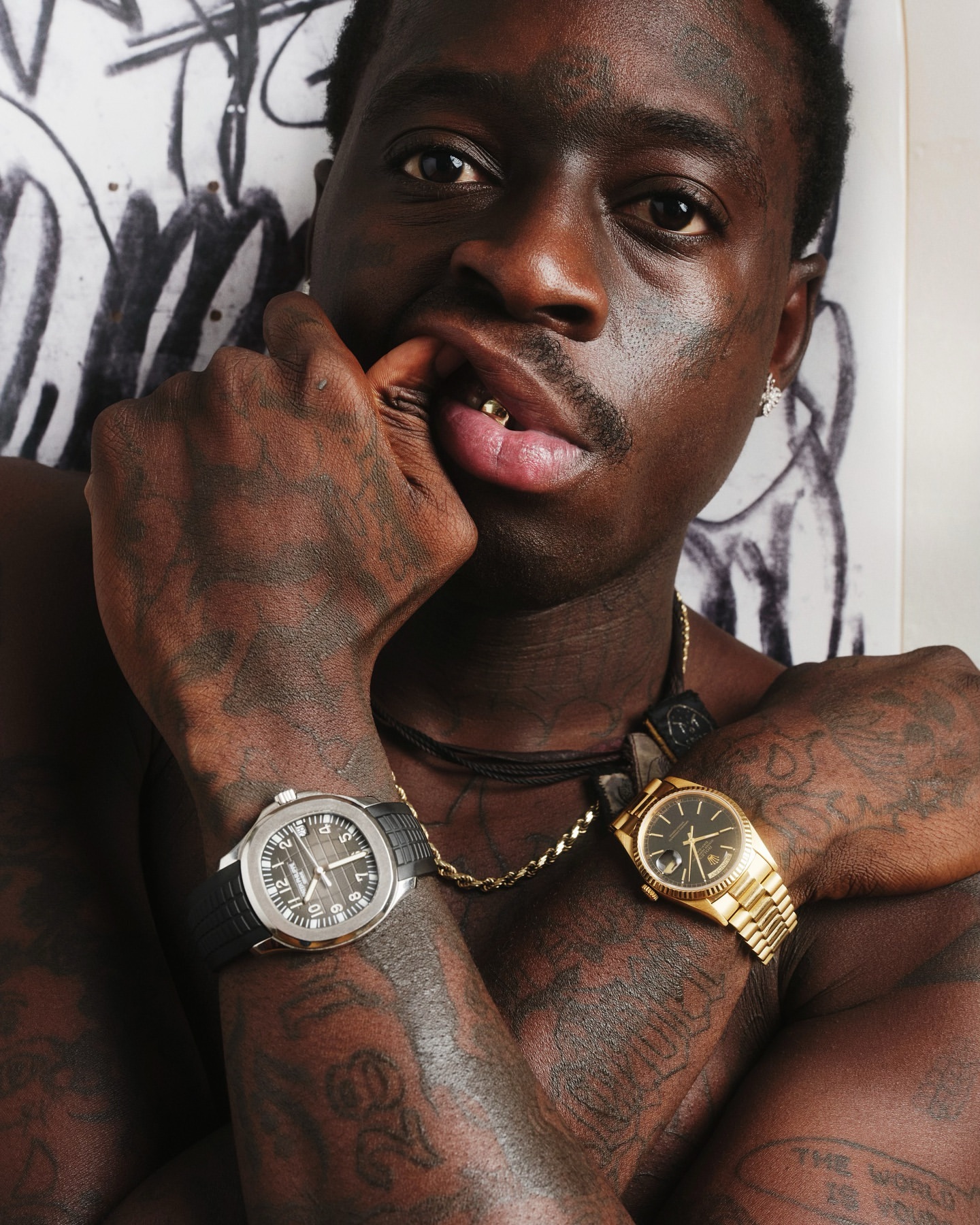 Close-up portrait of shirtless adult male with gold teeth, multiple watches and tattoos posing against a white graffiti background.