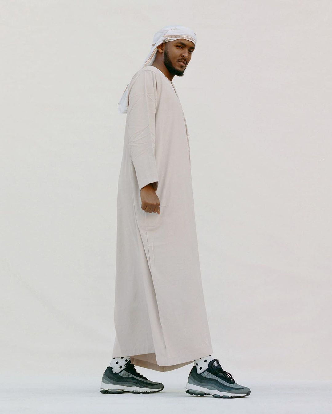 Adult male wearing traditional Emirati dress, including a white ghutra and kandura, with grey sneakers, standing against a light background.