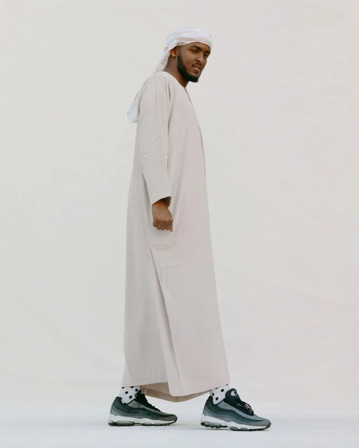 Adult male wearing traditional Emirati dress, including a white ghutra and kandura, with grey sneakers, standing against a light background.