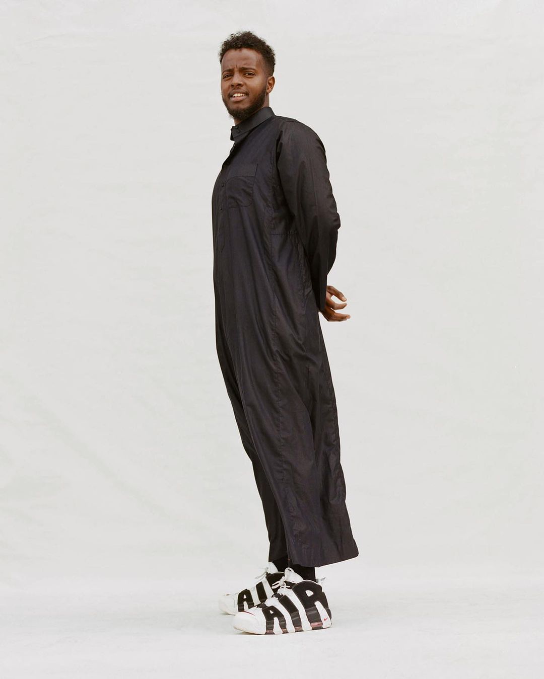 Portrait of a Black man in a long black coat and sneakers standing in a light studio. He has a slight smile and short curly hair.