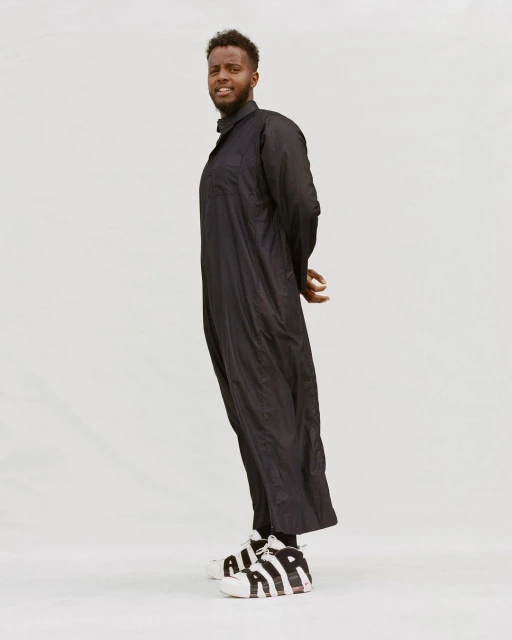 Portrait of a Black man in a long black coat and sneakers standing in a light studio. He has a slight smile and short curly hair.