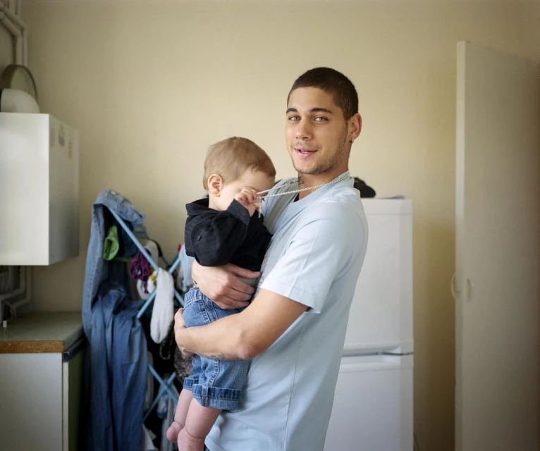 A young adult male holding a toddler in his arms indoors, with a neutral expression, medium shot, indoor setting, casual clothing, and soft lighting.