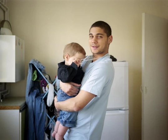 A young adult male holding a toddler in his arms indoors, with a neutral expression, medium shot, indoor setting, casual clothing, and soft lighting.