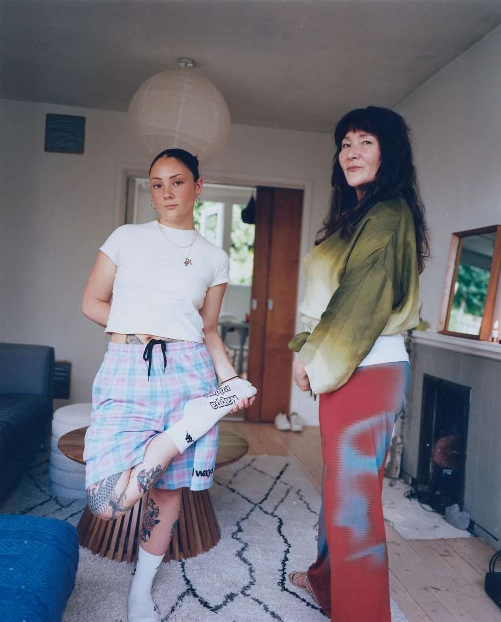 Two women pose indoors in casual clothes. One stands with a leg propped, showing a tattoo. The other stands beside her in a long skirt.