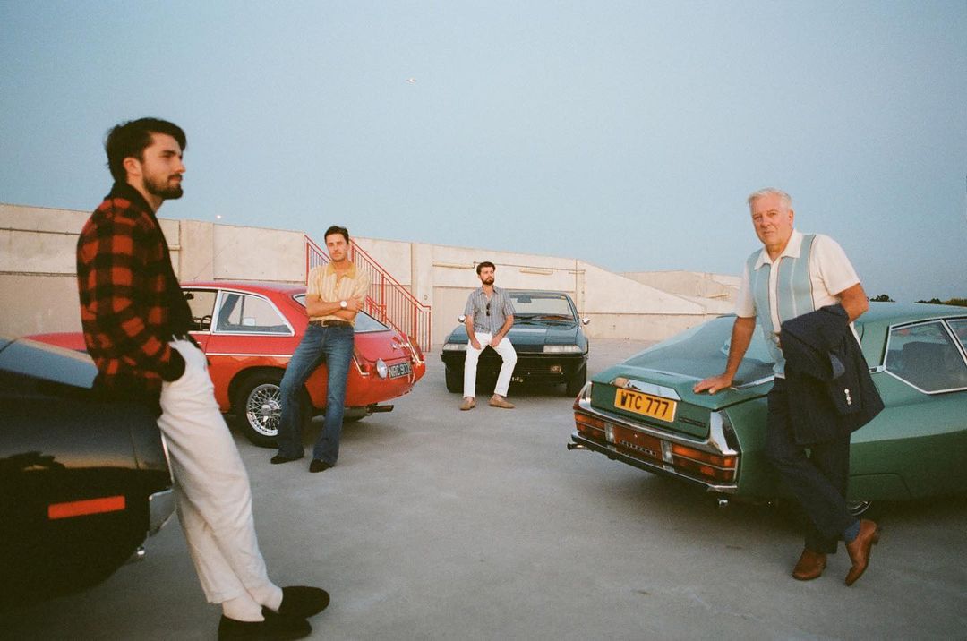 Group of four men pose with classic cars on a rooftop. One man leans on a black car, others pose by red, black, and green cars.