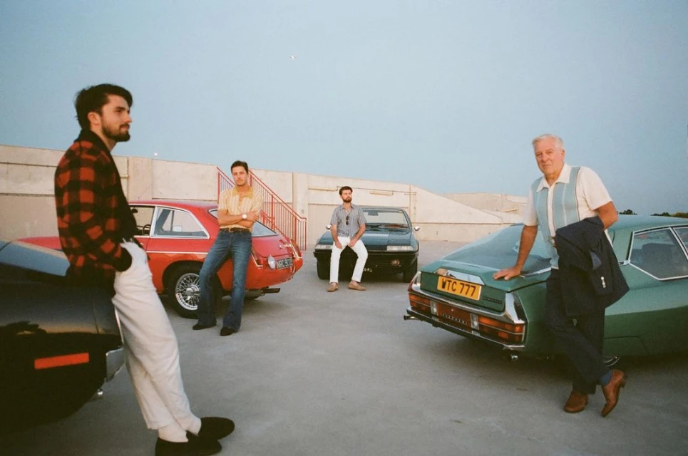 Group of four men pose with classic cars on a rooftop. One man leans on a black car, others pose by red, black, and green cars.