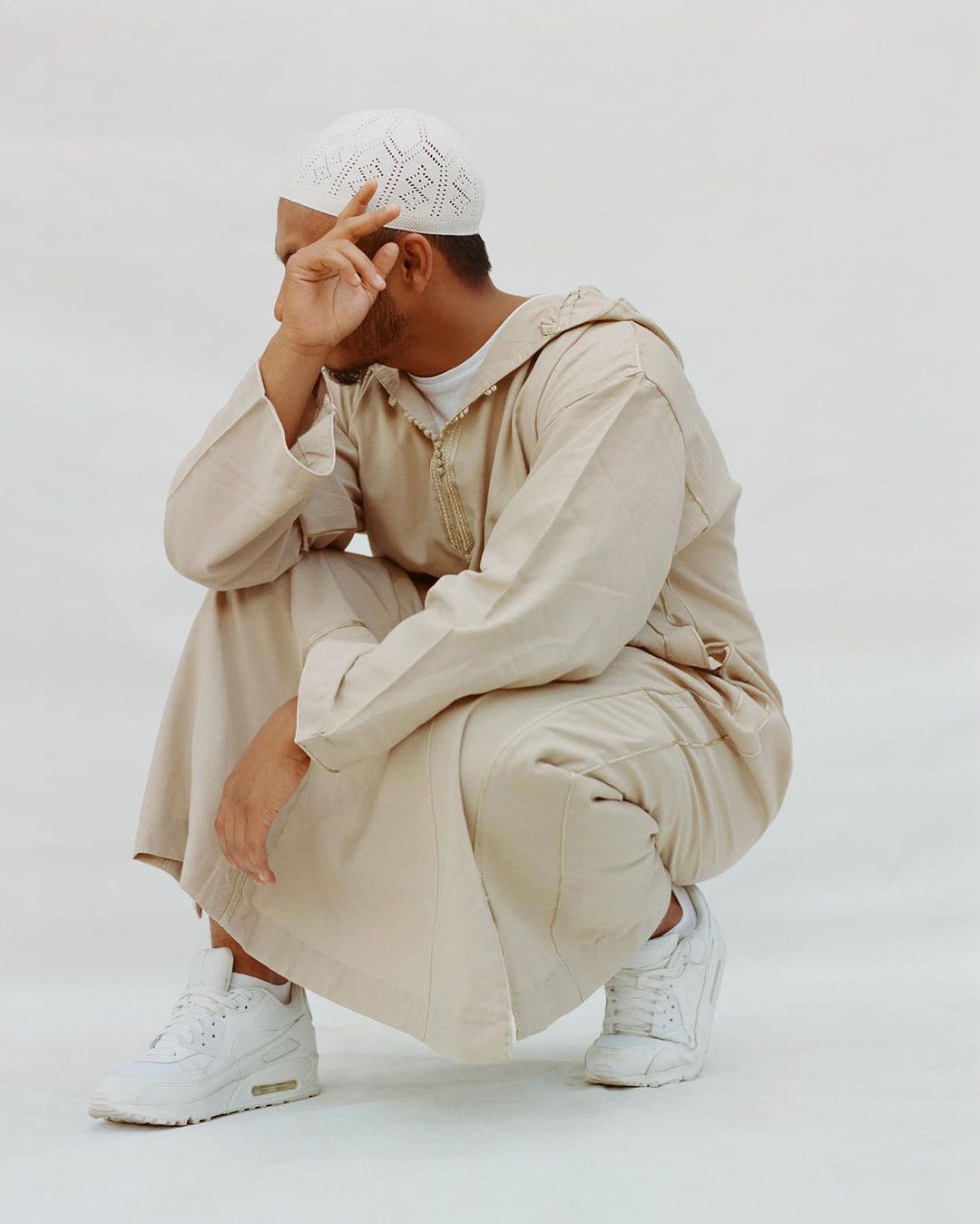 Man in traditional Islamic clothing squats against white background, hand near face, wearing white knit kufi and athletic shoes.
