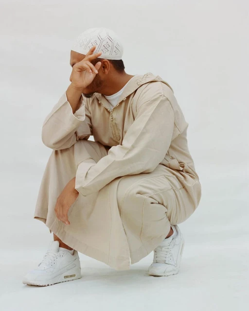Man in traditional Islamic clothing squats against white background, hand near face, wearing white knit kufi and athletic shoes.