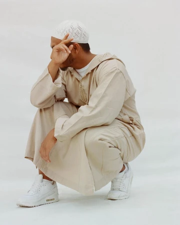 Man in traditional Islamic clothing squats against white background, hand near face, wearing white knit kufi and athletic shoes.