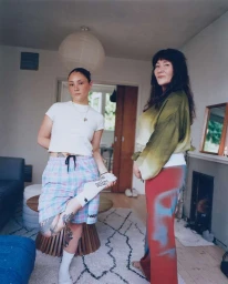 Two women standing in a living room, one holding her foot with white socks and plaid shorts, photographed by James Pearson-howes.
