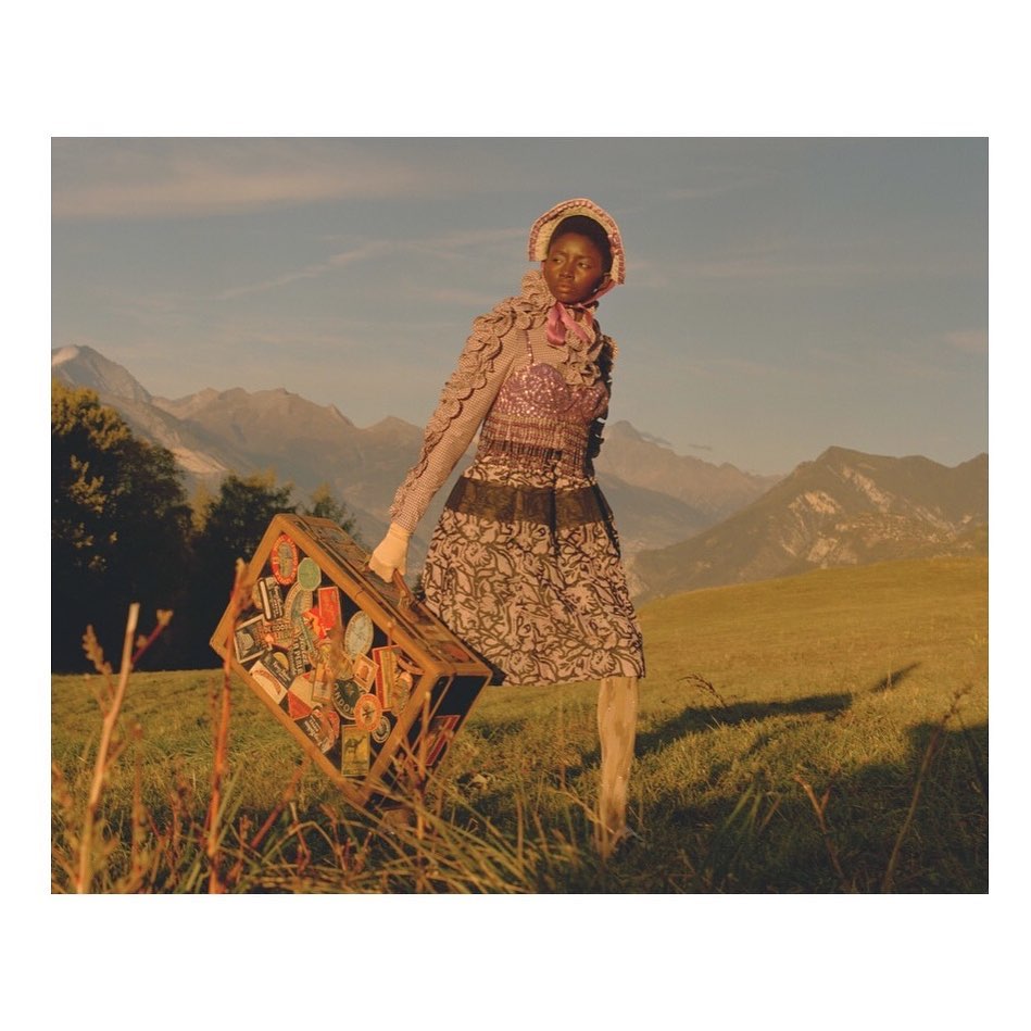 An adult female model carrying a suitcase in a grassy field with mountains in the background on a clear day in a fashion editorial.
