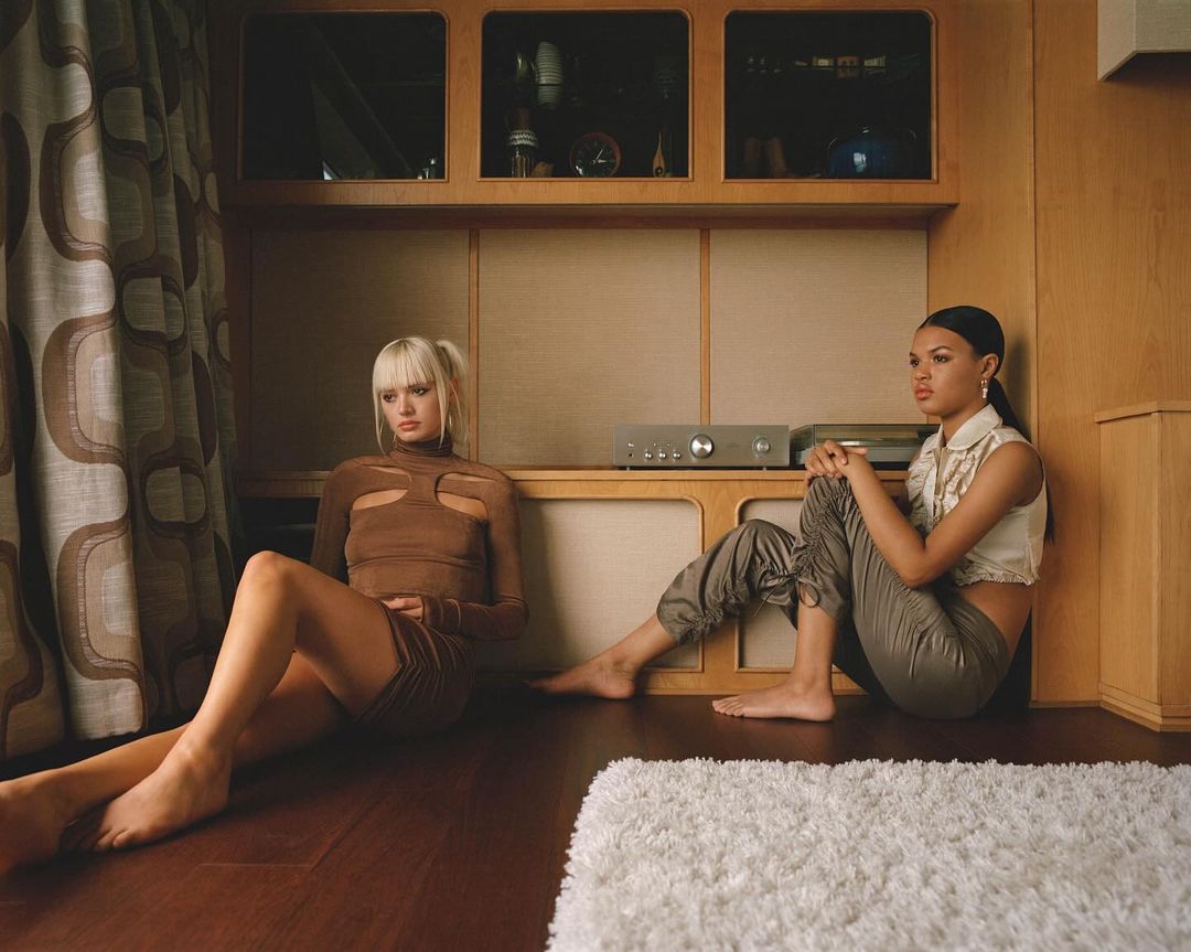 Two women in stylish outfits sit on a wood floor in front of a built-in cabinet and a white shag rug in an indoor setting.