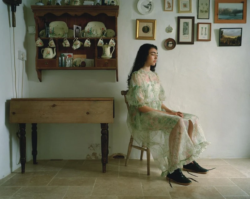 Woman in a floral dress sits in a room with a rustic cabinet, plates, and framed art, exuding a timeless, elegant atmosphere. 