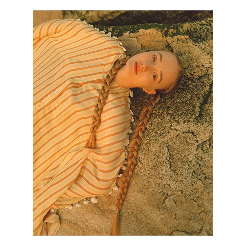 Eye-level shot of fair-skinned woman with blonde braids lying on a rock, wearing a tan and white striped shirt, portrait orientation.