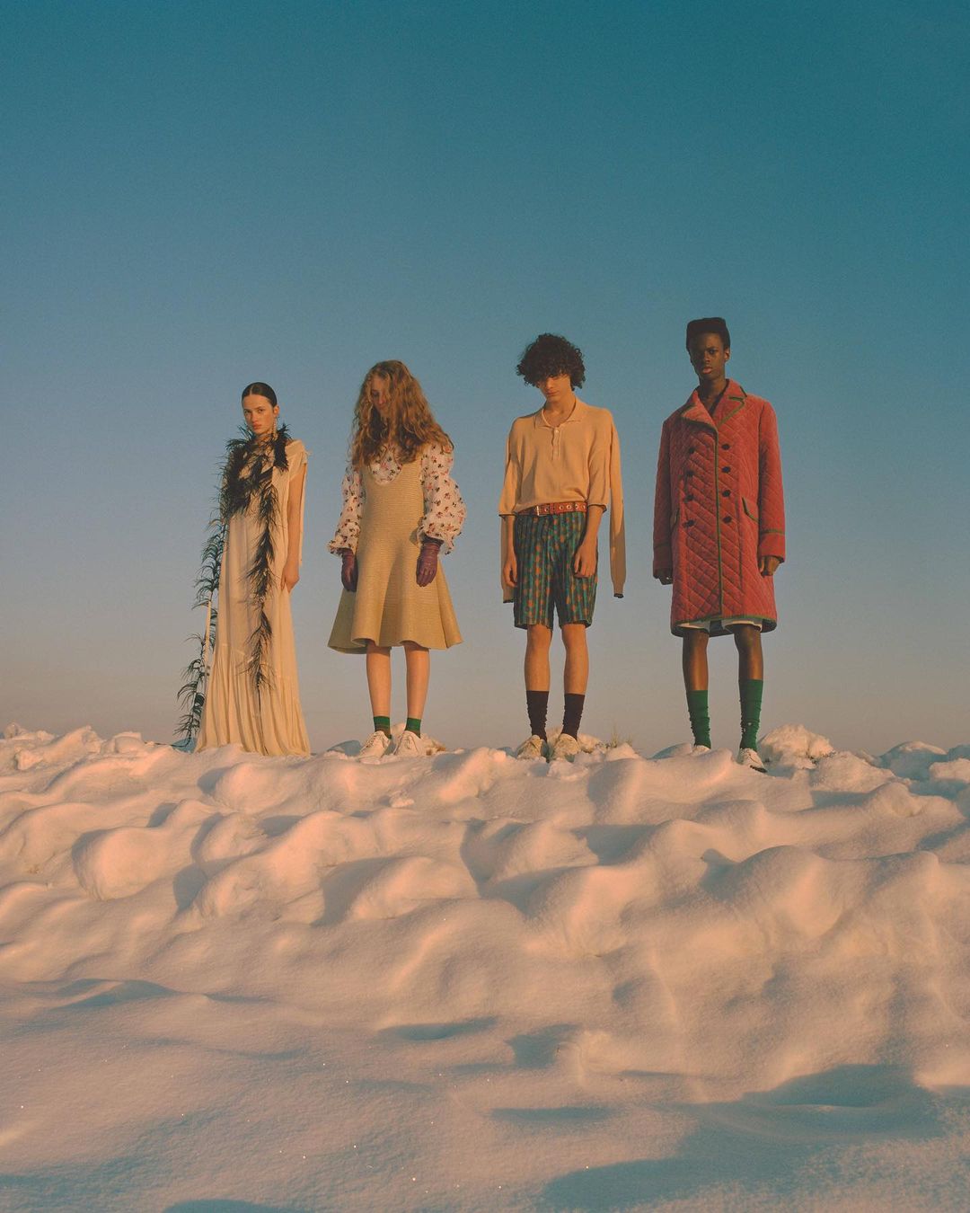 Four fashion models stand on a snowy hill under a clear sky. The models wear distinct outfits and pose for a fashion editorial.
