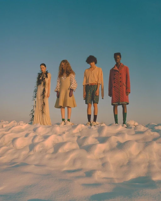 Four fashion models stand on a snowy hill under a clear sky. The models wear distinct outfits and pose for a fashion editorial.